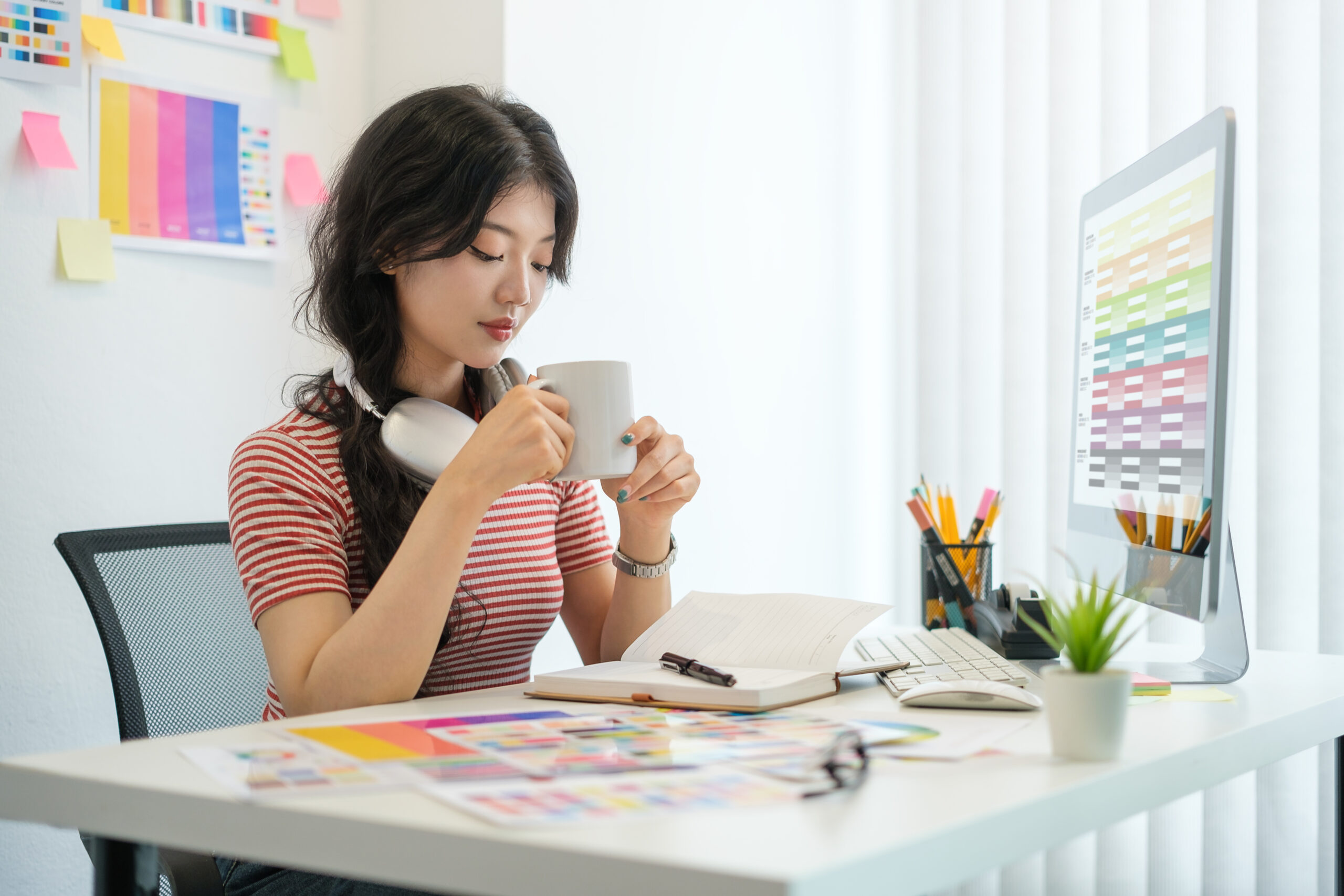 Image of satisfied young woman designer sitting at workplace and drinking coffee.